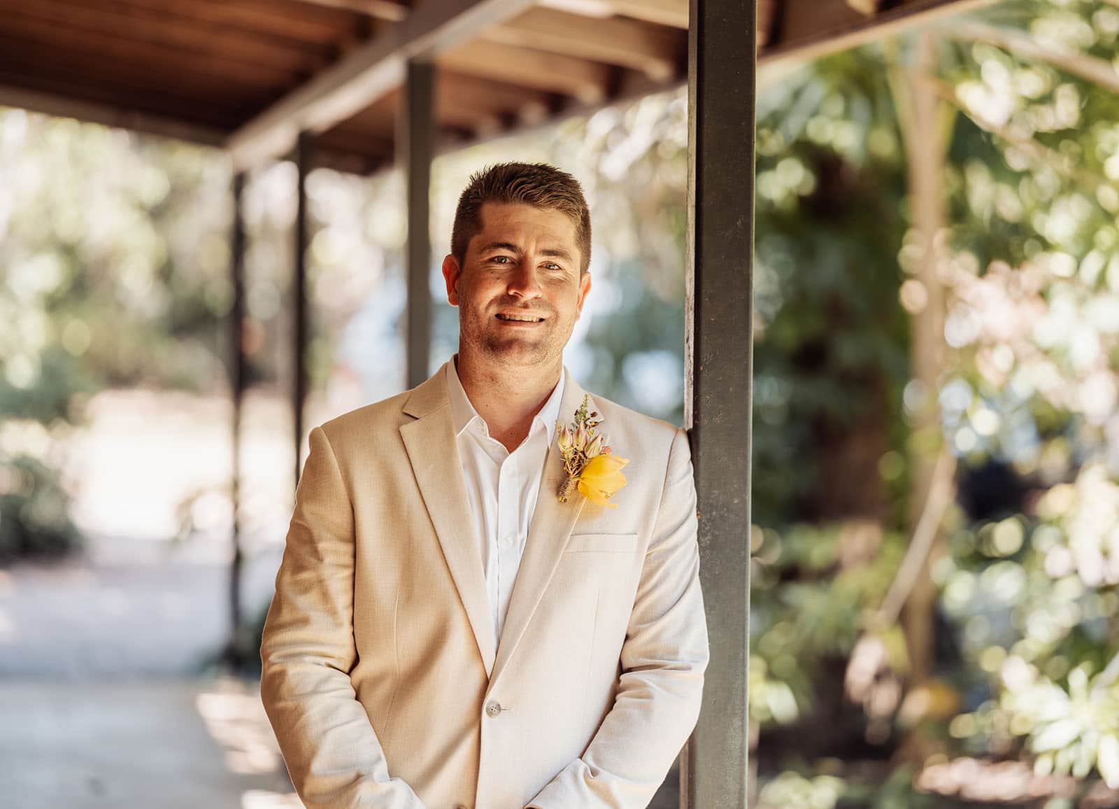 groom smiling under garden canopy Groom in beige suit, smiling under garden canopy with yellow boutonnière.